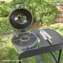 A round, grey charcoal grill with its lid open, set on a grassy lawn with shrubs in the background. The grill is mounted in the cart which has a side table, a metal grate inside, and a small warming rack on the left side of the grill. *Warming rack sold separately.