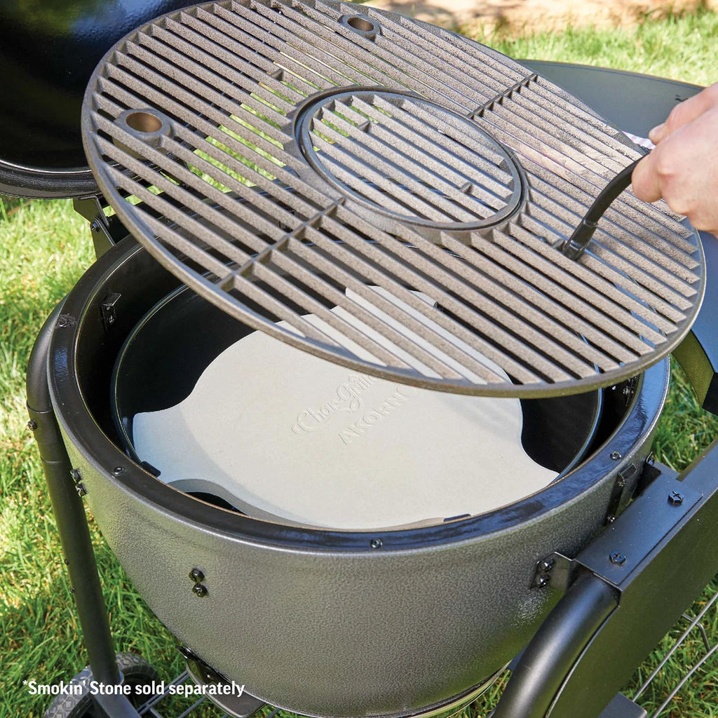 A close-up of a charcoal grill with its lid open, showing a round metal cooking grate being lifted by a hand using a grill gripper. Inside the grill is a white ceramic Smokin' Stone heat deflector.  *Smokin' Stone sold separately.