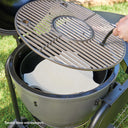 A close-up of a charcoal grill with its lid open, showing a round metal cooking grate being lifted by a hand using a grill gripper. Inside the grill is a white ceramic Smokin' Stone heat deflector.  *Smokin' Stone sold separately.