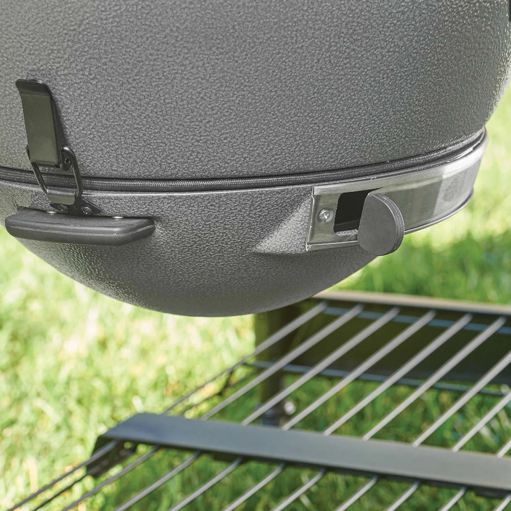 Close-up of a ash grey textured ceramic kamado with a metal draft door  and a latch holding the EasyDump Ash tray in place. Grill is positioned outdoors above the cart metal wire shelf with a blurred green grass background.