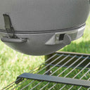 Close-up of a ash grey textured ceramic kamado with a metal draft door  and a latch holding the EasyDump Ash tray in place. Grill is positioned outdoors above the cart metal wire shelf with a blurred green grass background.