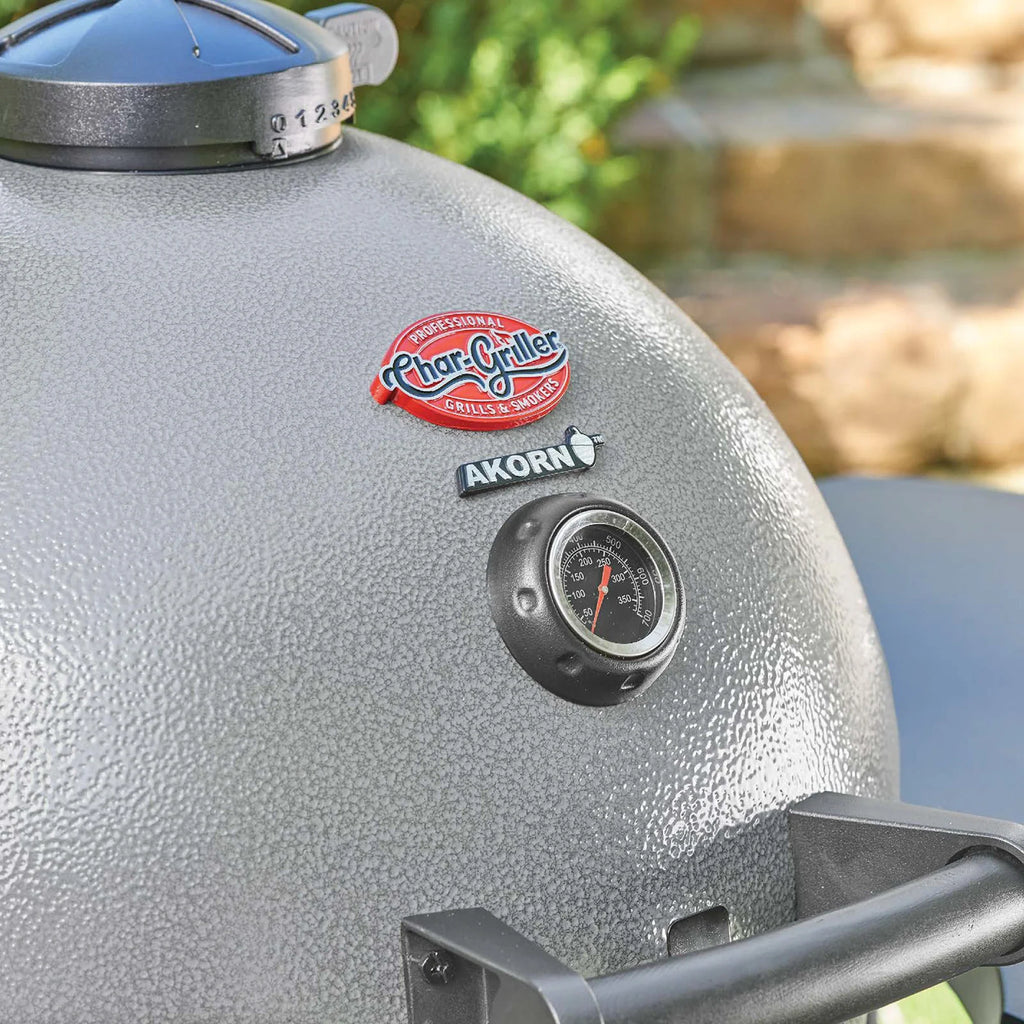 Close-up of a ash grey Char-Griller Akorn charcoal grill lid with a black dial thermometer and brand logo, set against a blurred green outdoor background.