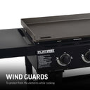 Closeup of half the griddle area showing a wall about 2 inches tall around side and back of griddle cooking surface. Text reads: wind guards to protect from the elements while cooking.