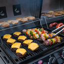 A wide variety of foods cook on both sides of the grill. On the gas side are cheeseburgers and kebabs with hamburger buns toasting on the warming rack. On the charcoal side there are sausages on the grill and potato wedges on the warming rack.