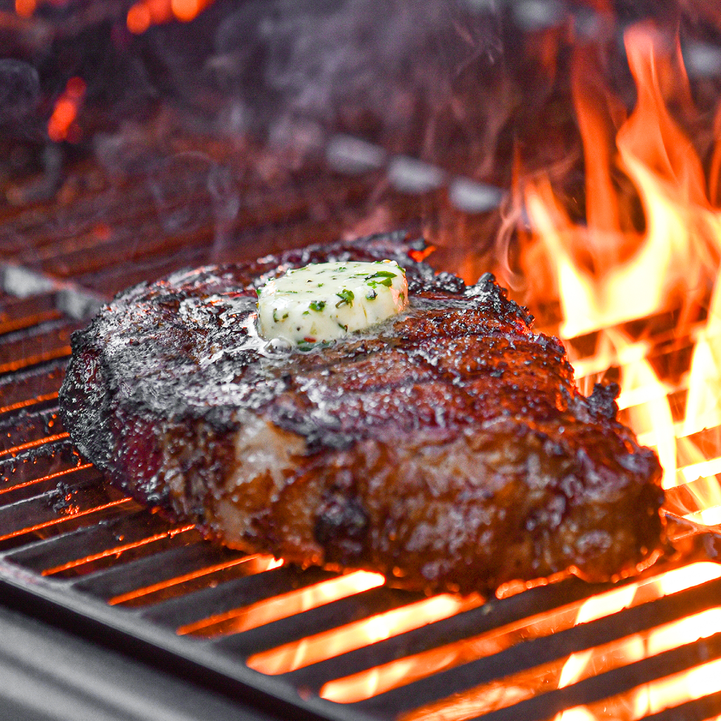 Flames shoot up through the cooking grate, searing a steak topped with a pat of compound butter