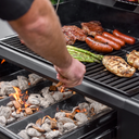 Steaks, sausages, chicken breasts and asparagus cook on the grill grate. A man adds wood chips over charcoal burning in the open Flavor Drawer