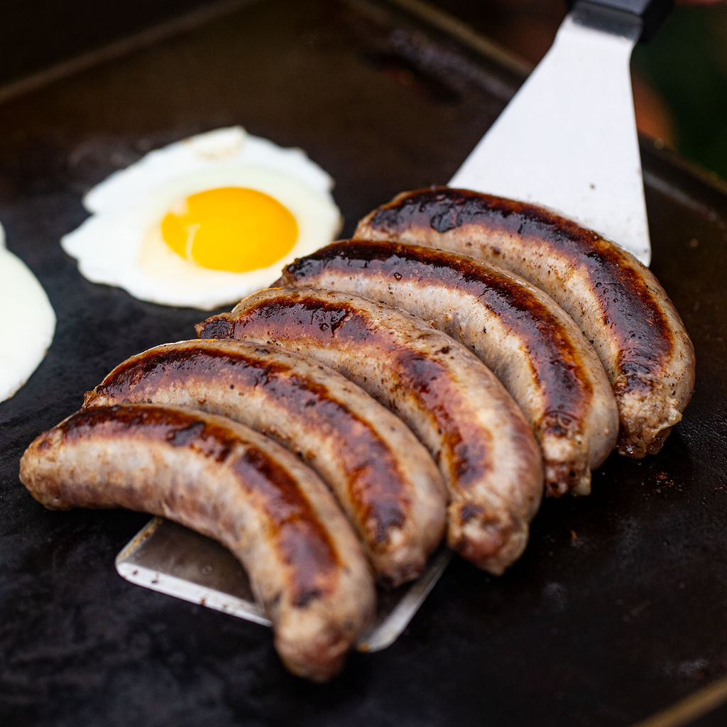 Someone uses a large spatula to remove 5 grilled sausages from the griddle. 2 eggs remain on the griddle.