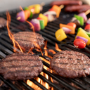 Flames lick up around 3 burgers on a grill cooking grate. Vegetable skewers are on the grates behind the burgers, and some sausages can be seen a bit further back.